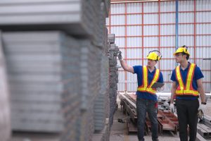Two workers wearing safety gear inspecting stacked steel materials inside a warehouse