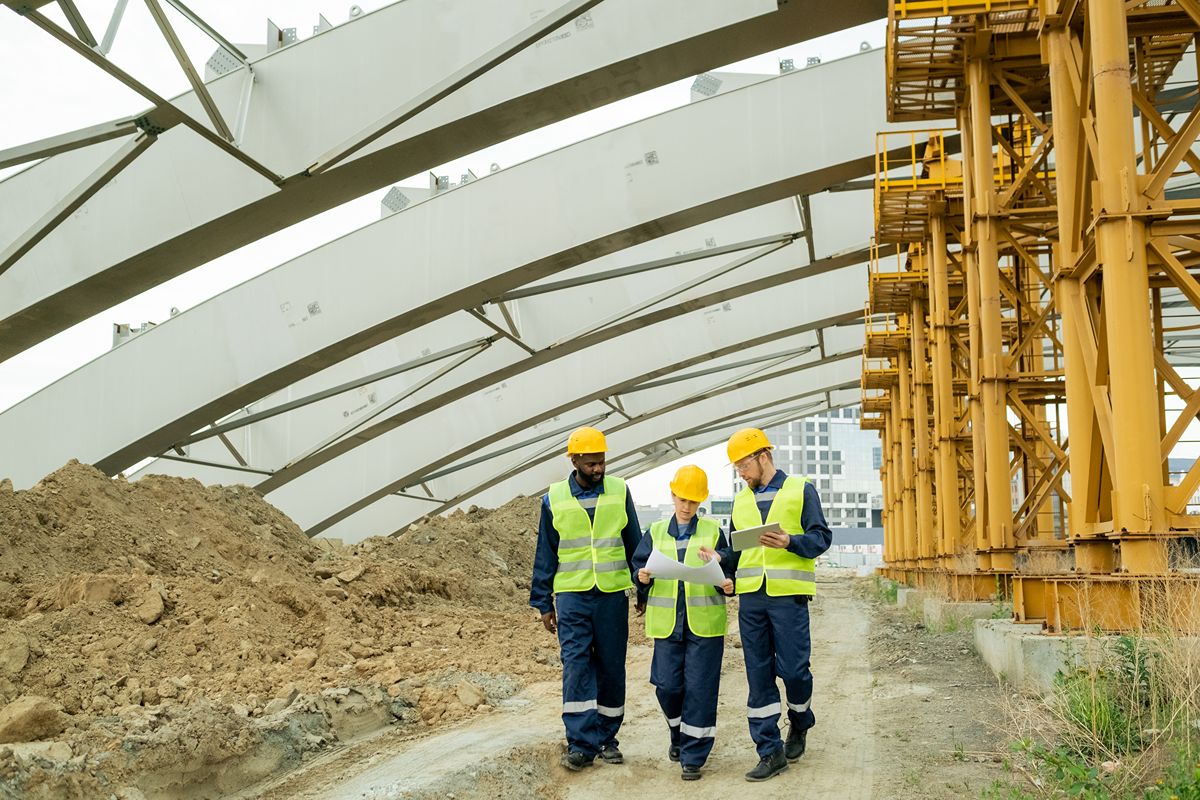 Three construction professionals reviewing plans beneath large steel beams at an active project site