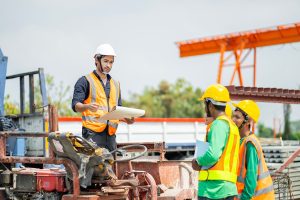 Site supervisor discussing plans with workers wearing safety gear near steel materials and heavy equipment